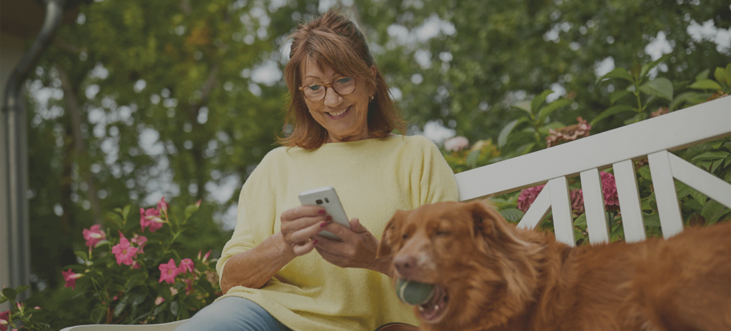 Eine Frau sitzt draußen auf einer Bank und schaut auf ein Handy. Neben ihr sitzt ein Hund.