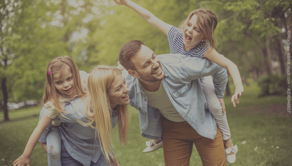 Eine Frau und ein Mann befinden sich in einer begrünten Anlage. Sie krümmen sich und tragen jeweils ein kleines Mädchen auf ihren Rücken.