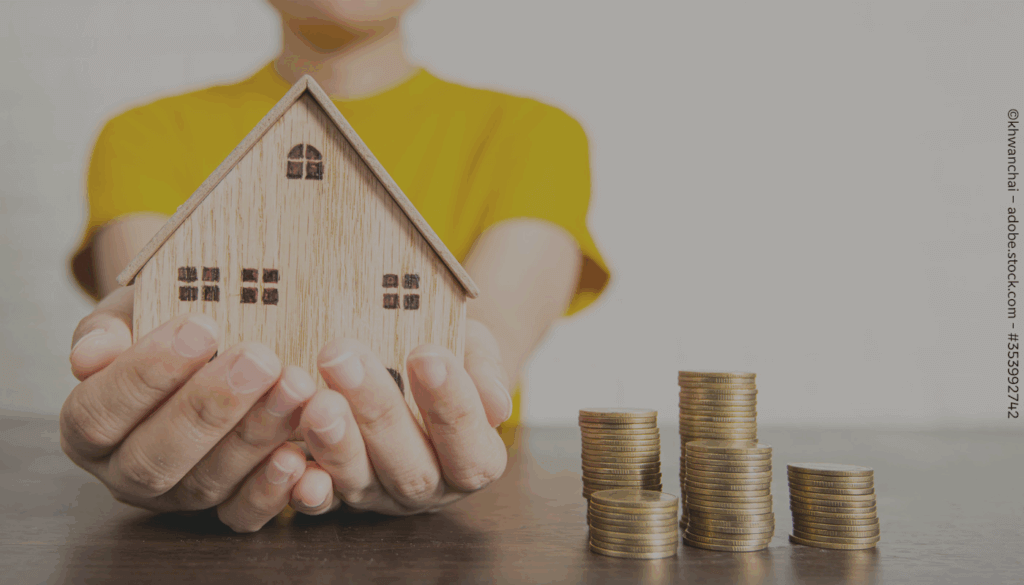 Eine Person hält ein Haus aus Holz in den Händen. Daneben sind drei Stapel mit goldenen Münzen auf einem Tisch.
