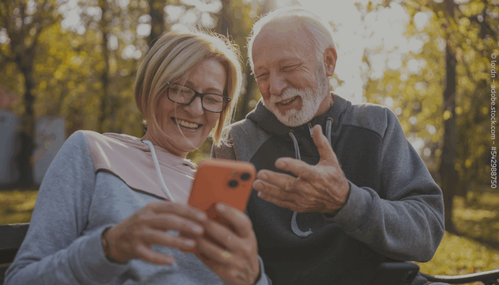 Eine ältere Frau und ein älterer Mann sitzen in einem Wald und schauen gemeinsam auf ein Smartphone, das die Frau in der Hand hält.