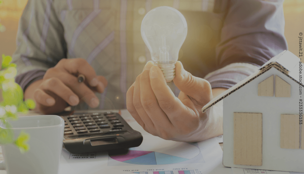 Eine leuchtende Glühbirne wird in einer Hand gehalten. Daneben liegt ein Taschenrechner und auf der anderen Seite steht ein Haus aus Pappe.