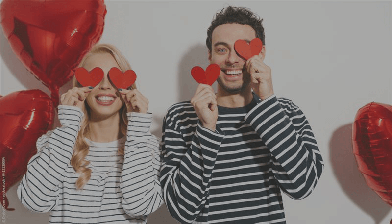 Eine Frau und ein Mann stehen nebeneinander und halten sich rote Herzen aus Papier vor das Gesicht. Neben ihnen sind Herzluftballons.