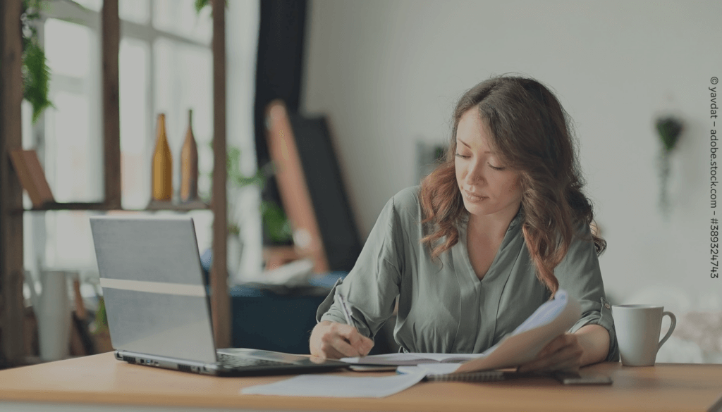 Eine Frau sitzt an einem Tisch vor einem Laptop und schreibt etwas in ein Heft.
