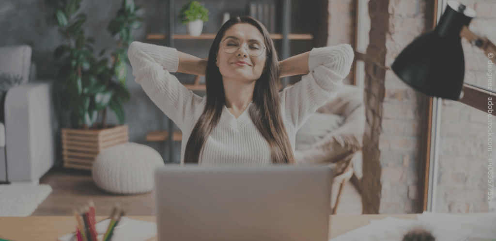 Eine Frau sitzt am Tisch vor einem Laptop und lehnt sich entspannt zurück.