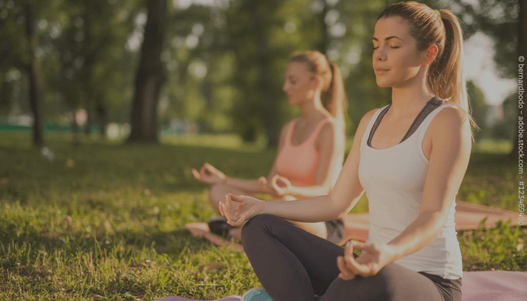 Zwei Frauen sitzen mit geschlossenen Augen auf dem Rasen in einer Yogapose.